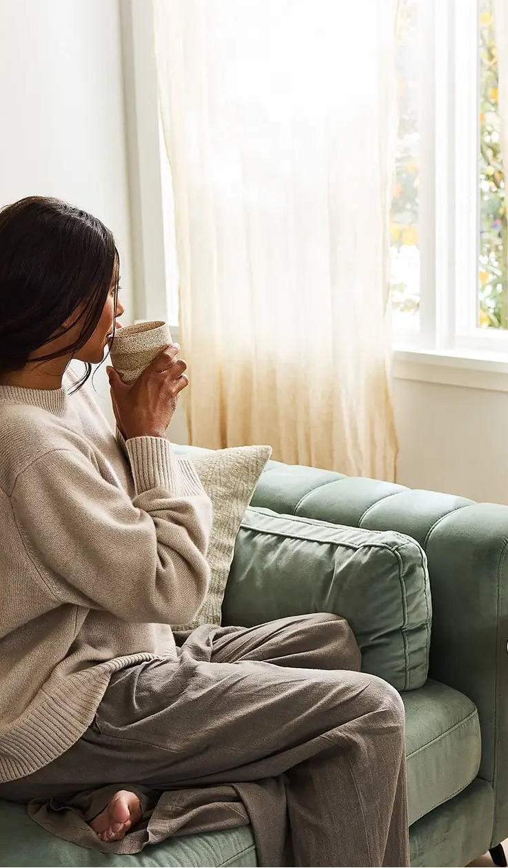 Woman sitting on a couch drinking coffee in a cozy living room.